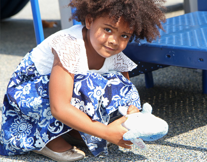 A young girl crouches and holds a stuffed whale toy on a playground.