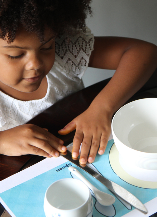 A young girl places a knife on a paper placement between a bowl and a spoon.