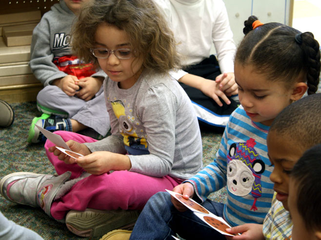 Two young girls sitting on a carpet hold paper cut-outs of cookies. Their classmates look on.