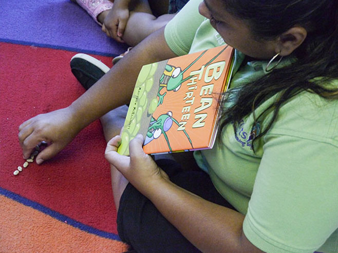 A teacher seated on the carpet shows the book cover and aligns dried beans in a row.