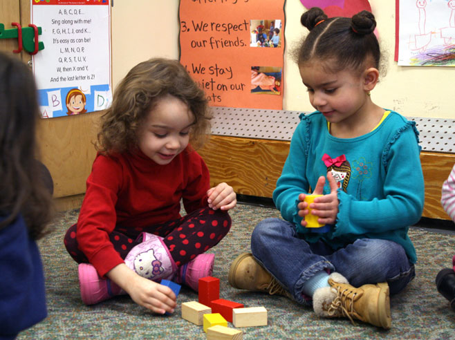 Two preschool girls sit on a carpet and take blocks from a pile.