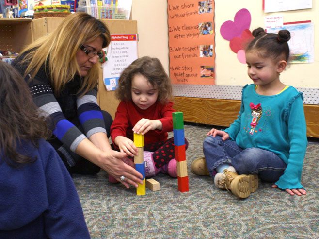 Two preschool girls sit on a carpet with block towers in front of them. One girl adds a block to her tower while the teacher holds it steady.