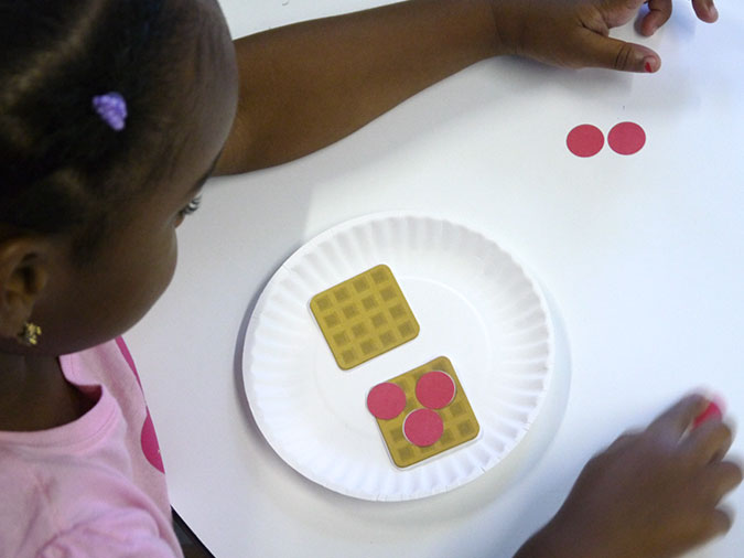 A young girl adds paper berries to her pretend waffles in a game of Breakfast Share.