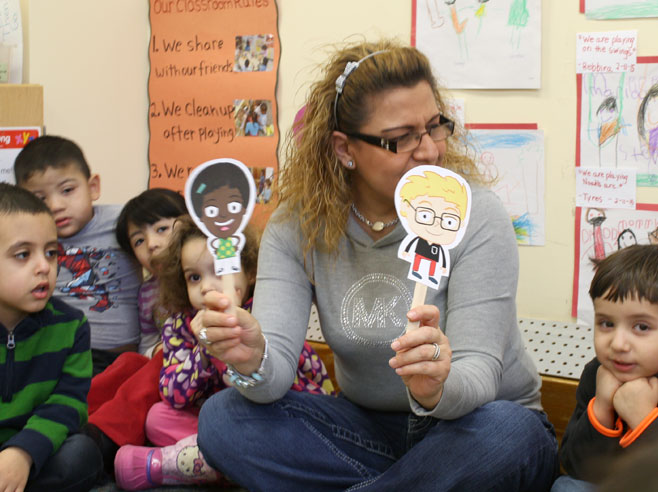A teacher sits on the floor in circle time and holds up paper cut-outs of two Gracie and Friends characters while children look on.