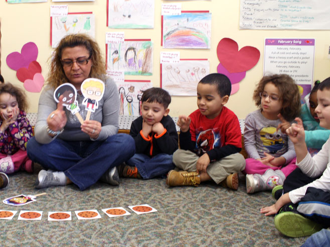 A preschool teacher shows paper cut-outs of two characters and six paper cookies. Children nearby point to count the cookies.