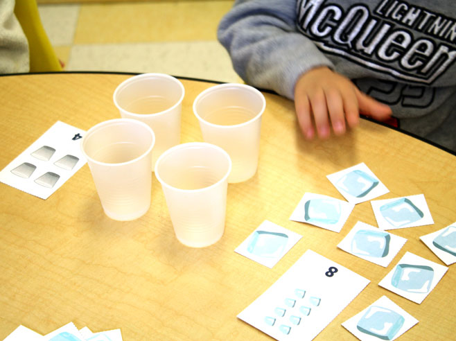 A preschool child in a gray sweatshirt reaches for the Drink Up Card Game materials on the table in front of him.