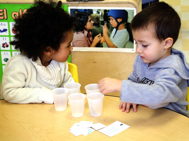 A boy in a grey sweatshirt sits at a table and puts paper ice cube cards into plastic cups as a girl in a white sweater watches.