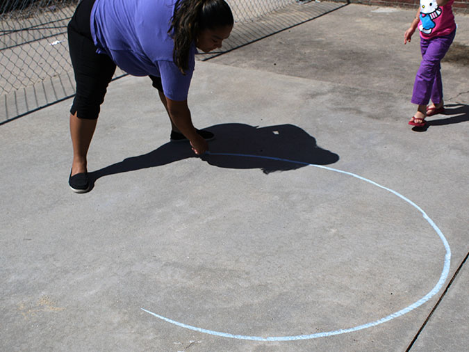 A teacher uses chalk to draw a circle on the playground pavement for Hopscotch Circle.