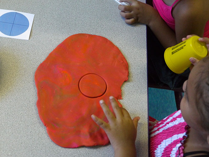 A preschool girl lifts the cup she used to cut a circle from a large flat piece of play dough.