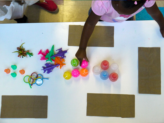 Small toys sorted by shape are in the center of a table with paper bags along the edges. A girl reaches for one of the toys.