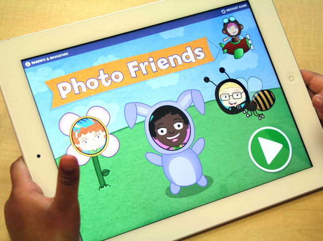 Close-up of a child’s hands holding an iPad with the Gracie and Friends Photo Friends app on-screen.