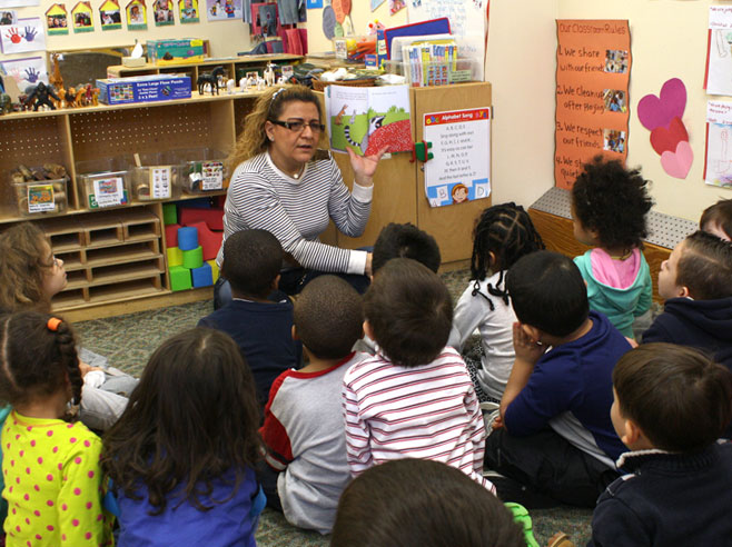 A teacher holds up the book and looks at the children in front of her to ask them a question.