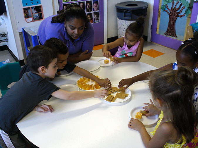 Children sit at a table set for snack and reach for crackers, orange slices, and cheese as their teacher looks on.