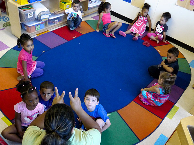 A preschool class sitting on the carpet watches their teacher hold up two fingers on each hand.