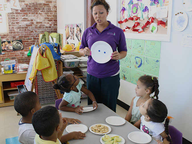 A teacher shows a dot plate to preschool children sitting at a table for snack.
