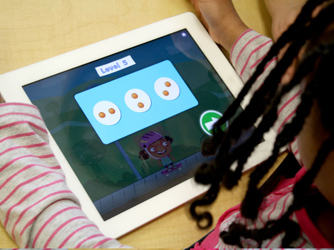 A preschool girl with braids looks at an iPad showing different arrangements of two oranges in a game level introduction.