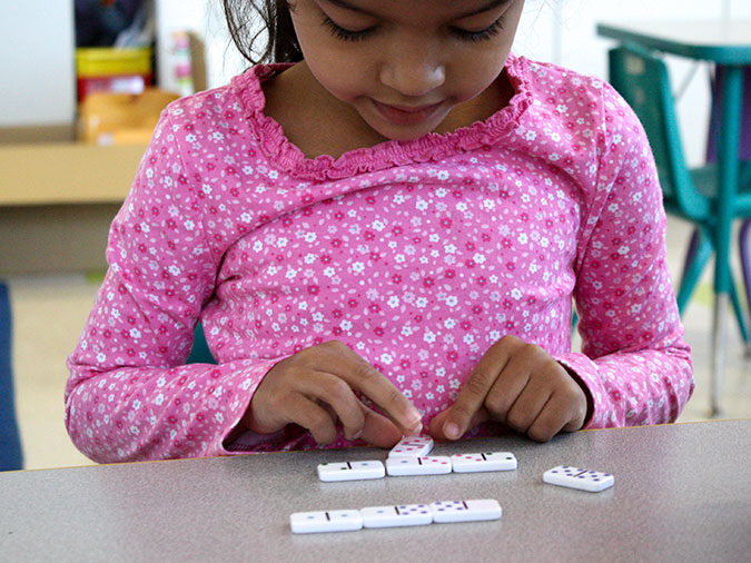 A girl in a pink shirt sits at a table and lines up dominoes with matching numbers.