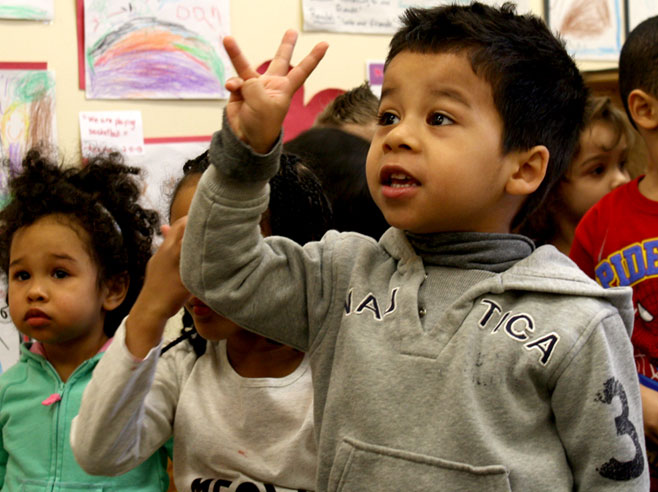A young boy in a grey sweatshirt, surrounded by classmates, holds up three fingers.