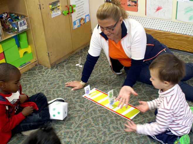 A preschool teacher and two children play the Jungle Gym Board Game on the carpeted floor.