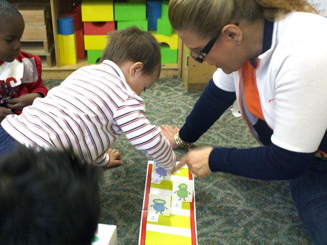 A teacher points to a space on the Jungle Gym Board Game. A young boy moves a purple robot game piece along the board.