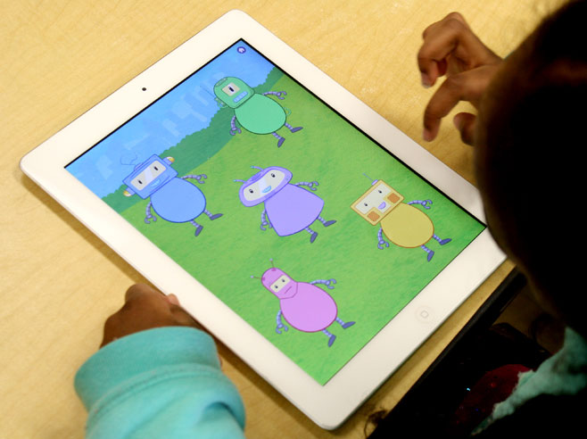 A young girl in a green sweatshirt looks at an iPad with five colorful robots on-screen.