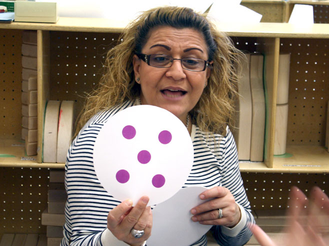 A teacher holds up a paper card with five purple dots on it.