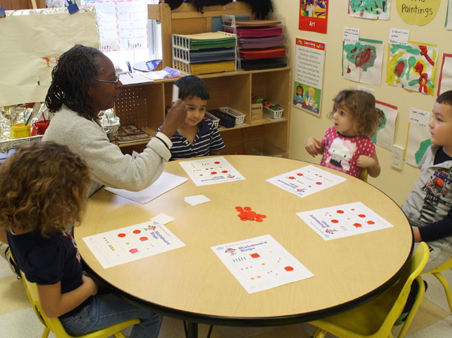 A teacher and four children sit at a table with board games and game tokens. The teacher holds up a small card.