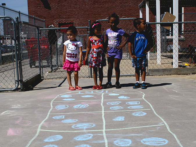 Four preschoolers stand on the playground in front of a double row of chalk boxes with two to four dots in each box.