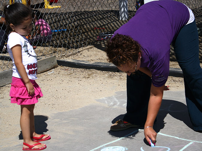 A young girl watches as her teacher draws dots in a square with chalk on the pavement.
