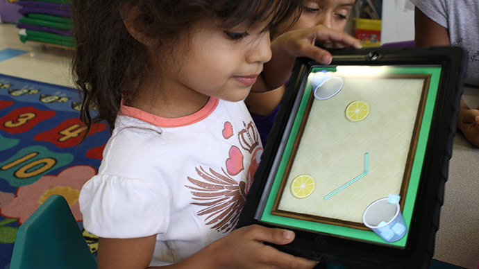 A preschool girl in a white and pink shirt plays the Early Math with Gracie & Friends Lemonade Stand app by tilting her iPad and watching the ice cubes in the app slide into cups.