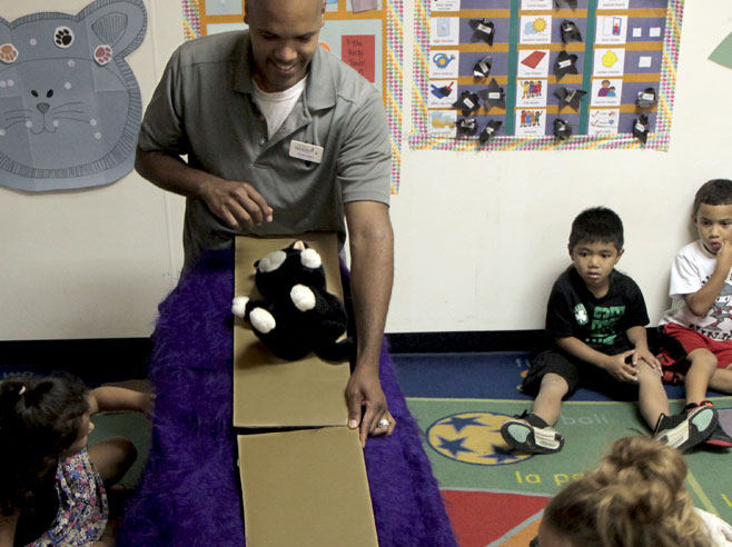 As students watch, a teacher slides a stuffed animal down a makeshift cardboard ramp.