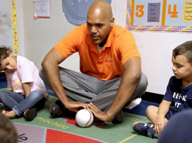 A teacher, sitting with students in circle time, gets ready to push a baseball across the floor.