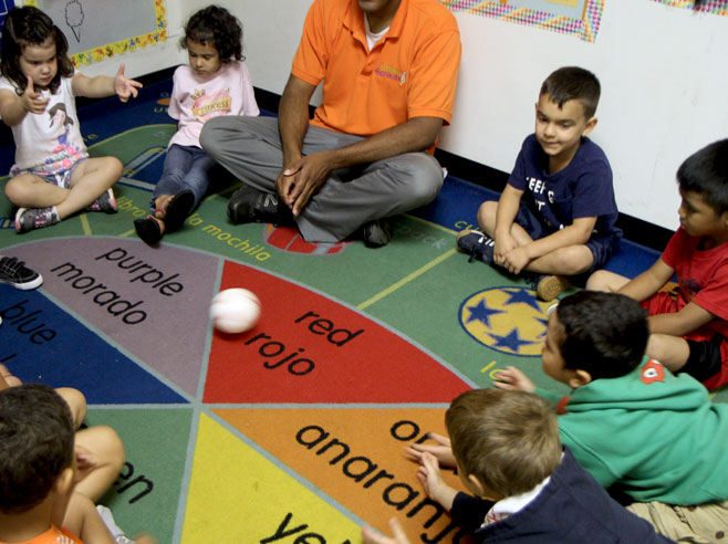 Students and a teacher sit in circle time and watch a ball roll across the floor.