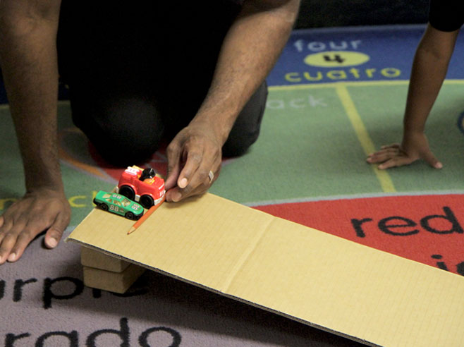 A teacher's hands can be seen holding a pencil to keep two toy cars from going down a cardboard ramp.