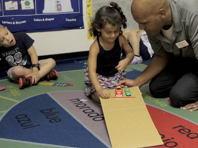 A student and teacher kneel over a gently sloping cardboard ramp. The student holds a pencil in front of two toy cars, to keep them from going down the ramp.
