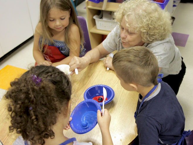 At a table, students and a teacher have bowls, with paint and a giant marble in each. One girl tilts the bowl to roll the marble around in the paint.