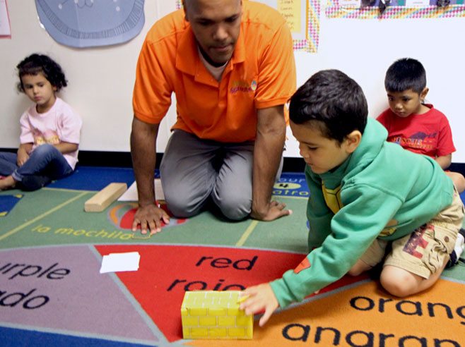 A teacher and students look on, as a boy pushes a cardboard block across the rug.