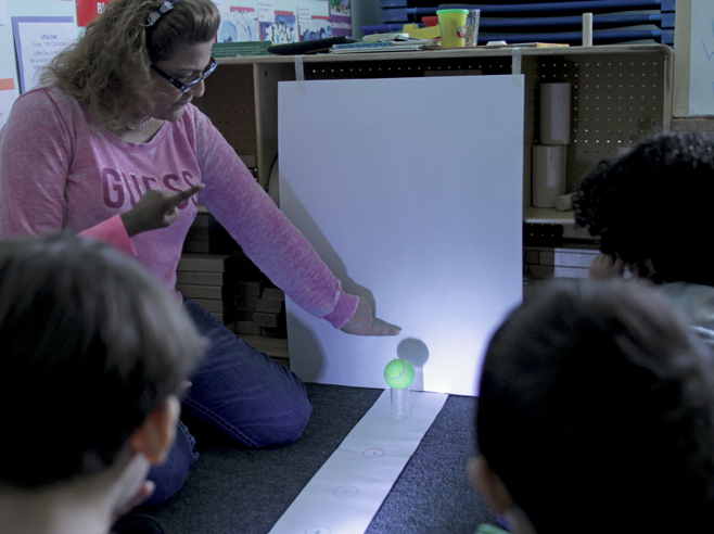 A teacher draws with crayon on a posterboard around a shadow, made by light shining at a ball on a cup.