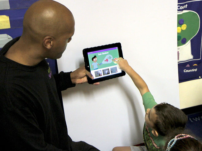 Teacher holds up iPad showing Cool Shadows app screenshot. A student points at the screen.