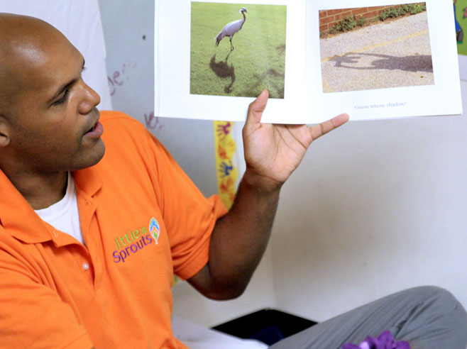 A teacher holds a book up. The book is open to show some photos of shadows, one created by a bird, and the other created by a child holding up a stuffed animal.