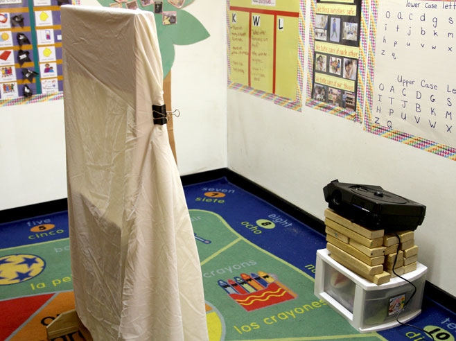 A classroom puppet stage sits in the middle of the room. A large white sheet is draped over it and attached with binder clips. Behind it, a slideshow projector can be seen, stacked up on wooden blocks.