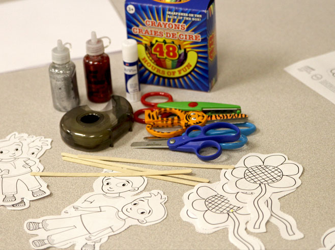Materials for making shadow puppets are laid out on the table: crayons, glue, glitter, scissors, character cut-outs, tape, and craft sticks.
