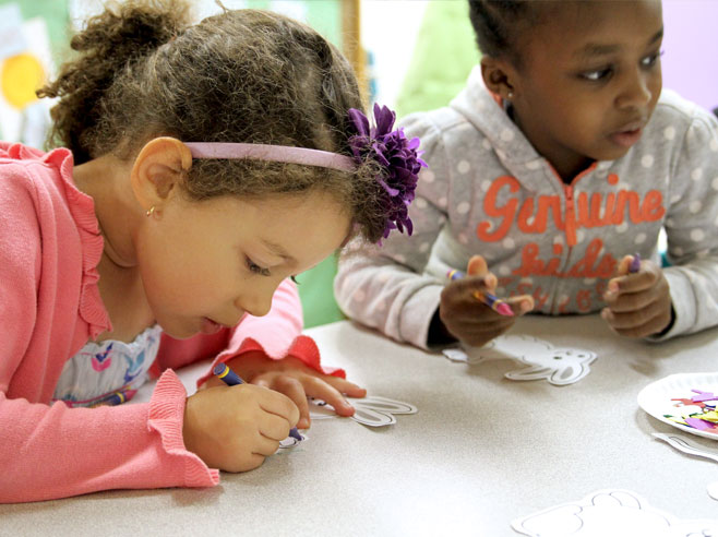 Two students sit at a table and use crayons to color their paper character cut-outs.