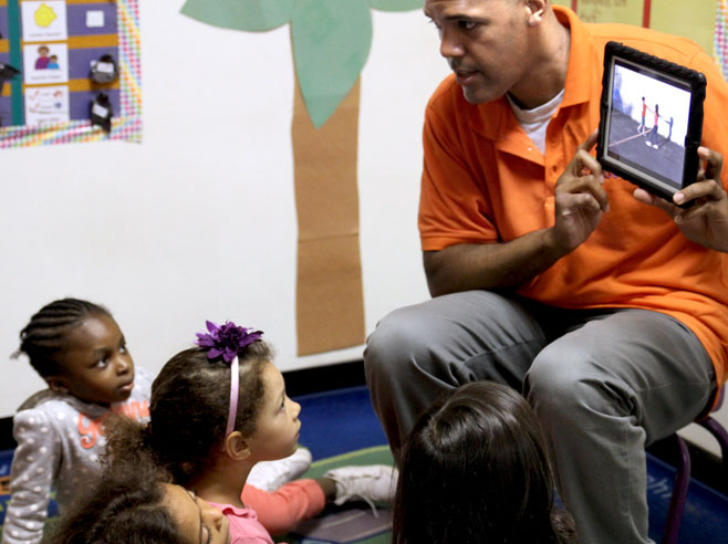 A teacher sits on a chair in front of students, who are seated on the floor. He is showing a video on an iPad.
