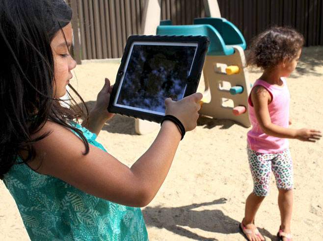 A student holds an iPad up in a sandy playground. She is taking a picture of another student, running with her shadow clearly visible.