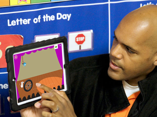 Teacher holds up iPad, and points at screen showing a Shadow Play app game screenshot.