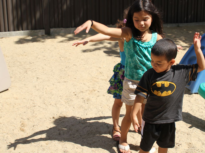 Three students stand one behind the other, in a sandy playground. They wave their arms to create interesting shadows on the ground.