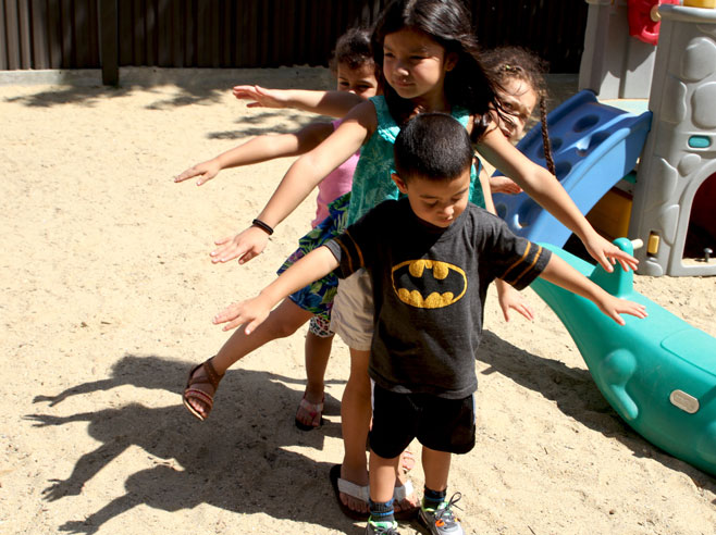 Four students stand one behind the other, in a sandy playground. They wave their arms to create interesting shadows on the ground.