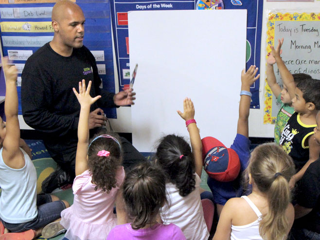 A teacher kneels in front of a group of students. There is a poster-board against the wall, and he holds a shadow puppet in one hand, and a flashlight in the other.