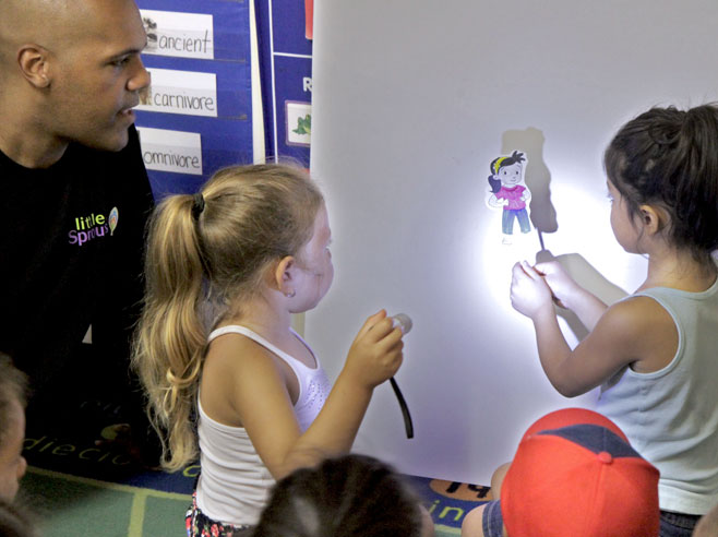 One student holds a hand-made shadow puppet (a cut-out of a PBS character on a craft stick) against a piece of white poster-board on the wall. Another student shines a flashlight at it, creating a shadow.